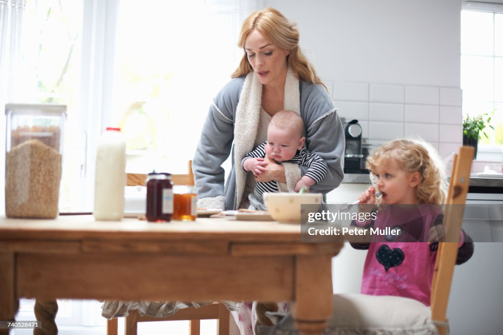Mother holding baby boy, sitting at kitchen table with young daughter, having breakfast