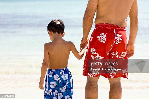 rear view of a boy holding his father's hands standing on the beach - swimming shorts stock pictures, royalty-free photos & images
