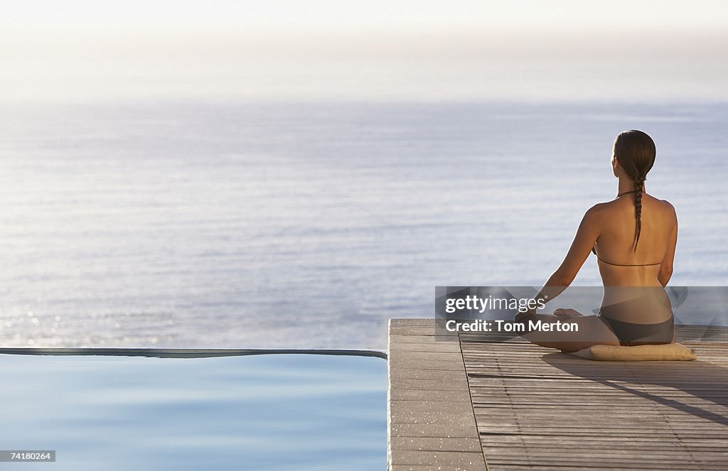 Woman sitting on infinity pool deck doing yoga rear view