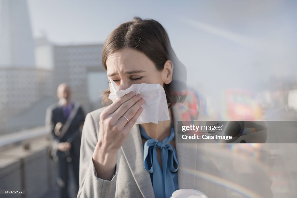 Businesswoman with allergies blowing nose into tissue on sunny urban sidewalk