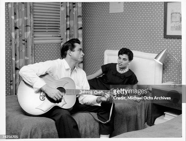 Country singer/songwriter Johnny Cash sits on a bed playing acoustic guitar as his first wife Vivian Liberto looks on in circa 1957.