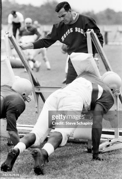 View of University of Notre Dame football coach Ara Parseghian as he oversees his players practice tackling using a football sled on the field, South...