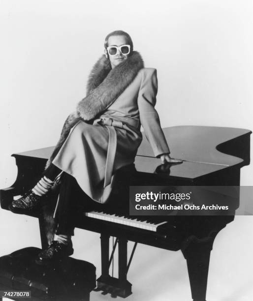 Pop singer Elton John poses for a portrait sitting on top of a piano wearing a fur-lined coat in circa 1974.