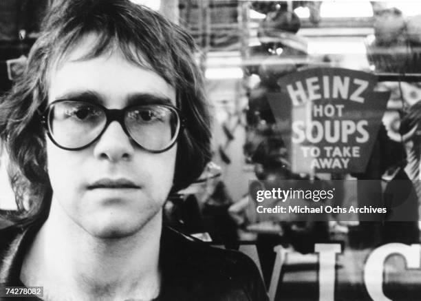 Pop singer Elton John poses for a portrait wearing glasses in front of a sign that says "Heinz Hot Soups" in circa 1970.