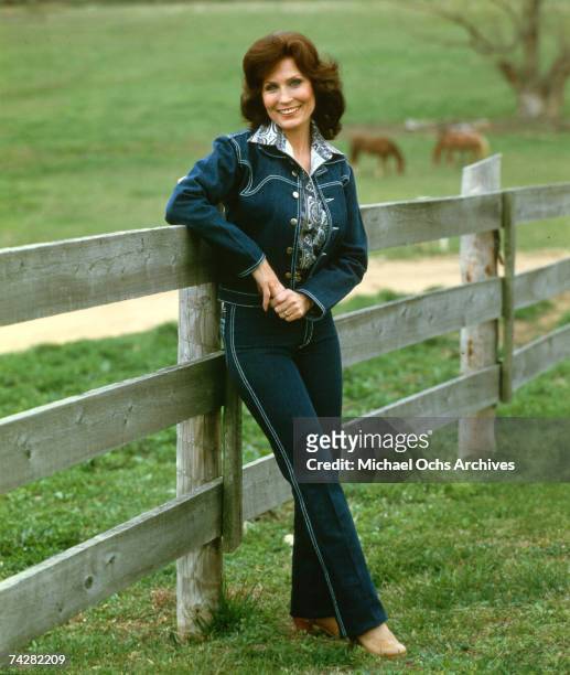 Loretta Lynn poses for a portrait wearing a blue denim suit with cows in the background leaning up against a fence in circa 1972.