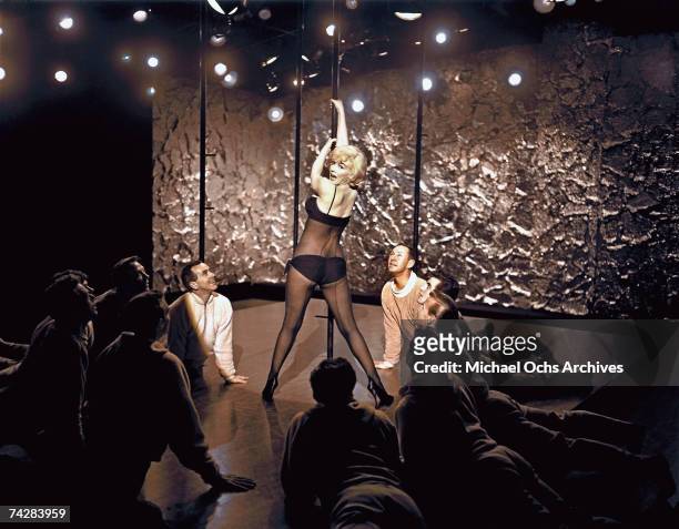 Actress Marilyn Monroe poses for a portrait hanging on a stripper pole wearing lingerie surrounded by a cadre of actors ogling her in a scene from...