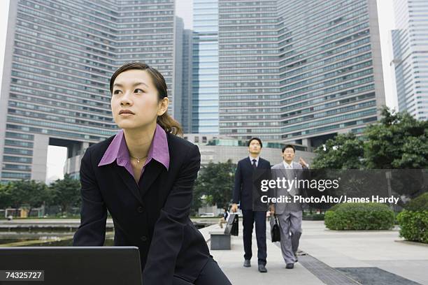 businesswoman using laptop in office park, two businessmen approaching in background - gap closers stock pictures, royalty-free photos & images