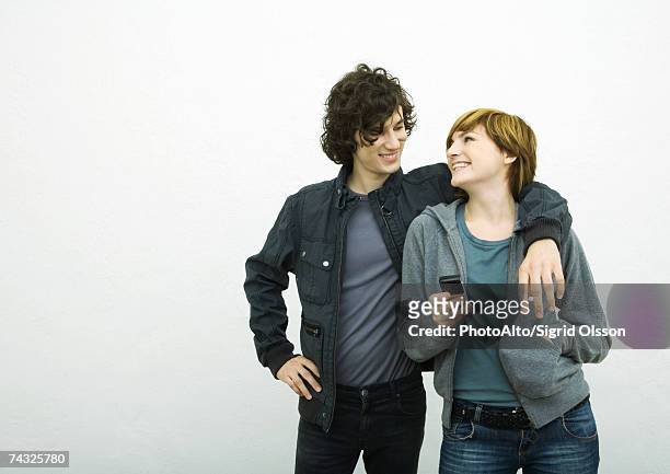 young adult couple looking at each other, man with arm around woman, woman holding cell phone - couple studio shot stock pictures, royalty-free photos & images