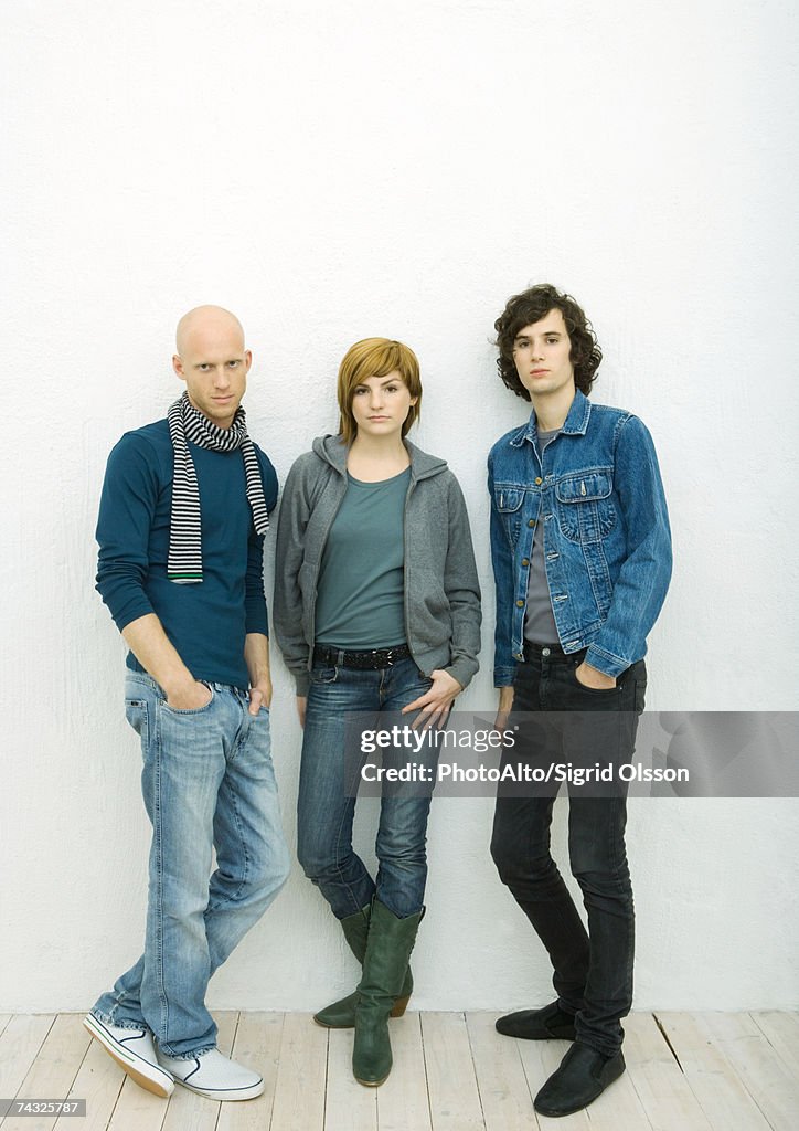 Three young adults leaning against wall, looking at camera, full length portrait, white background