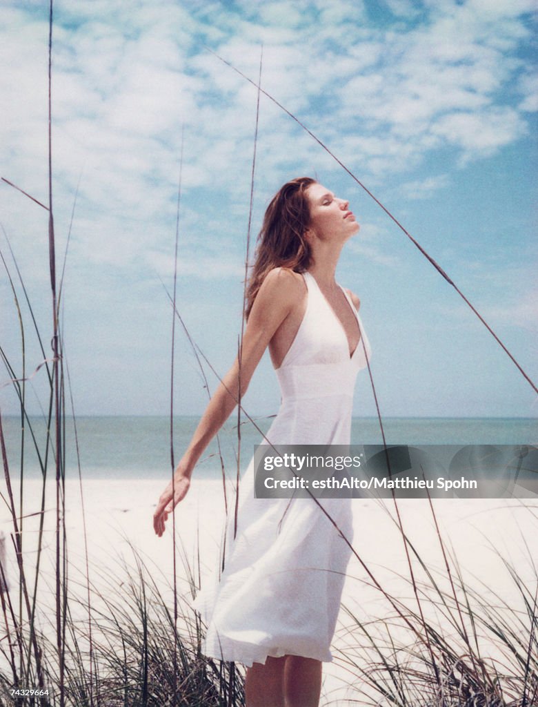 Young woman in dress standing in dune grass with head back, facing wind