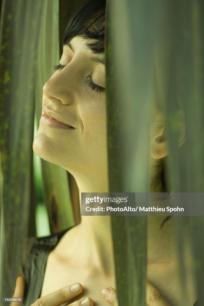 Woman standing among palm leaves, eyes closed, hands on chest