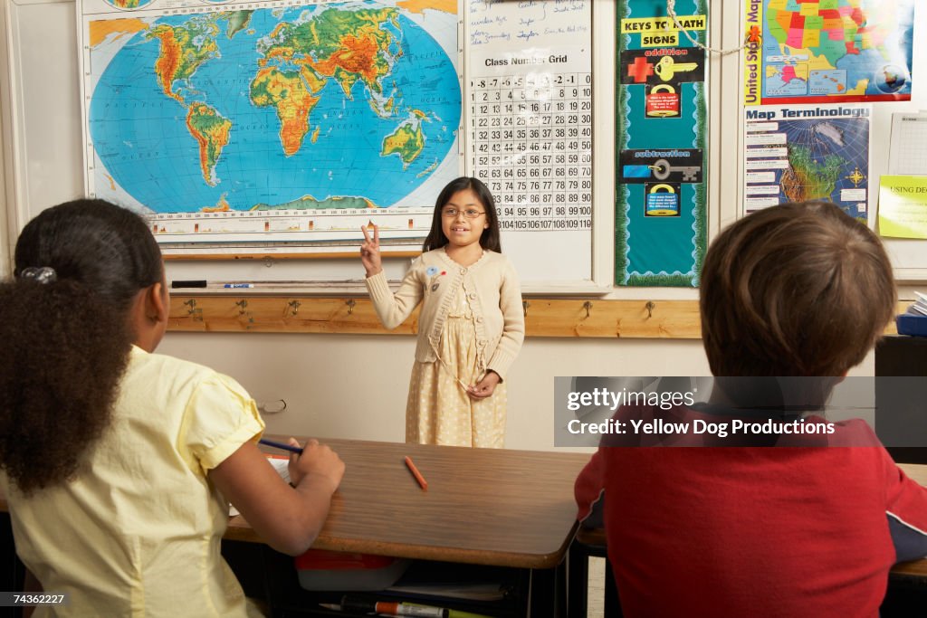 Girl (8-9) demonstrating peace sign in front of classroom, other students watching