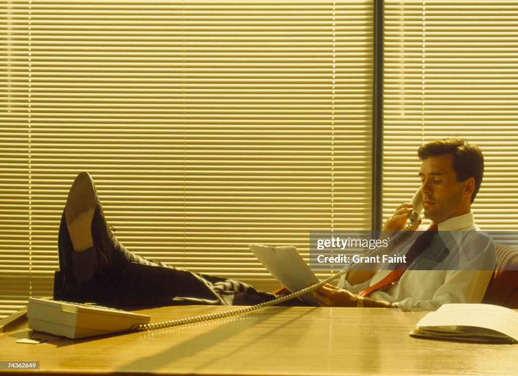 Mid adult businessman in office, sitting at desk with feet up, using phone