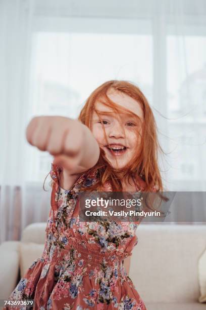 portrait of cheerful girl punching while standing against sofa in living room - faustschlag stock-fotos und bilder
