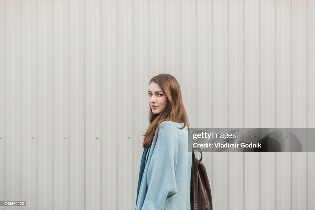 Portrait of beautiful young woman carrying backpack while standing by wall