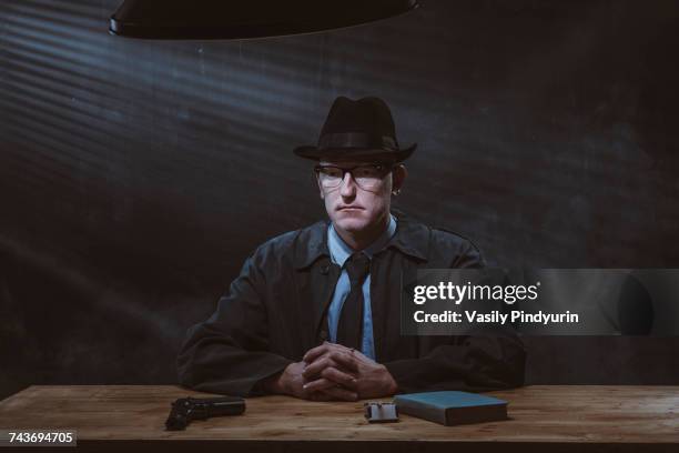 portrait of young man sitting at table with gun against black wall - investigatore foto e immagini stock