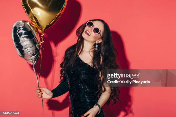 happy woman holding helium balloons against coral background - black sequin dress stock-fotos und bilder