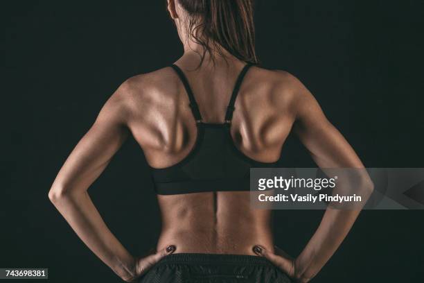 rear view of female athlete wearing sports bra standing with hands on hip against black background - struttura muscolare del torso foto e immagini stock