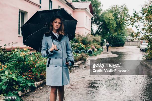 portrait of teenage girl holding umbrella while standing on wet footpath by buildings in rain - russia rain boots imagens e fotografias de stock