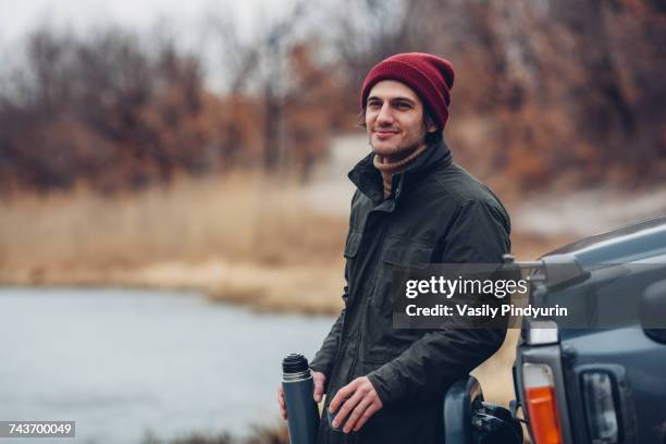 smiling man holding insulated drink container while standing by sports utility vehicle at lakeshore - insulated drink container foto e immagini stock