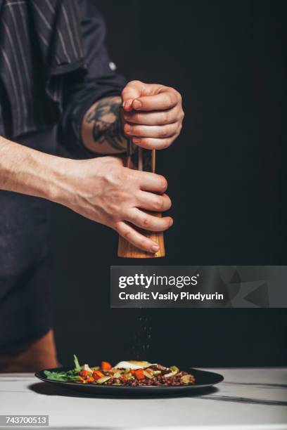 cropped image of man sprinkling from pepper mill over food on table against black background - pepper mill stockfoto's en -beelden