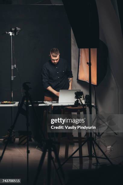 Photographer working on laptop while standing by food plate against backdrop at studio