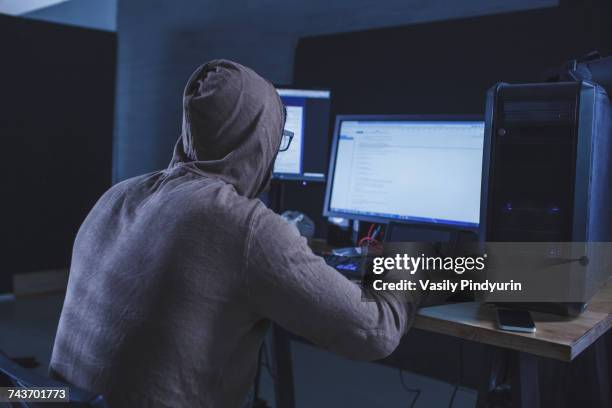 computer hacker wearing hooded shirt using computer at table - anonymous hacker stockfoto's en -beelden