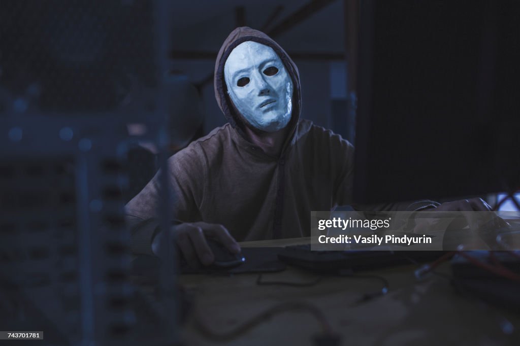 Computer hacker wearing mask and hood using computer while sitting at table
