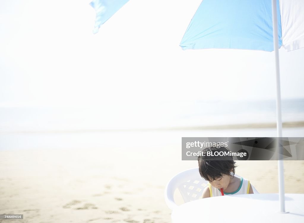 Japanese boy sitting on chair under parasol on beach