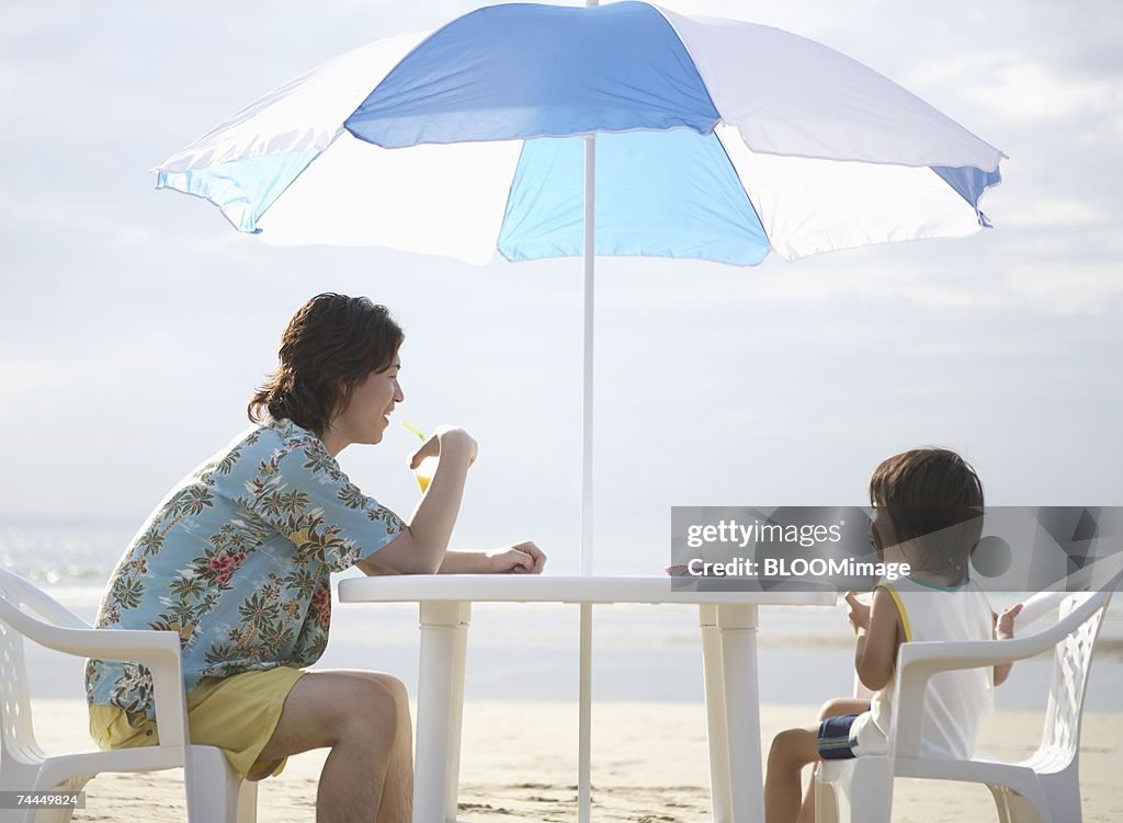 Japanese man and boy sitting on chair under parasol on beach