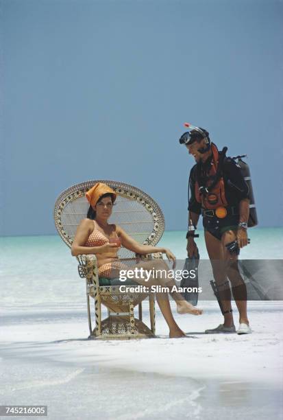 Man wearing diving equipment talks with a girl in a wicker chair on the beach, Bahamas, 1967.