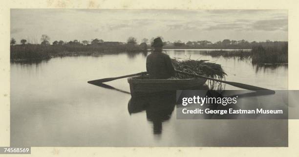 Entitled 'Rowing Home the Schoof-Stuff' , the image shows a man as he rows a skiff with bundles of reeds on a river, England, mid 1880s. The images...