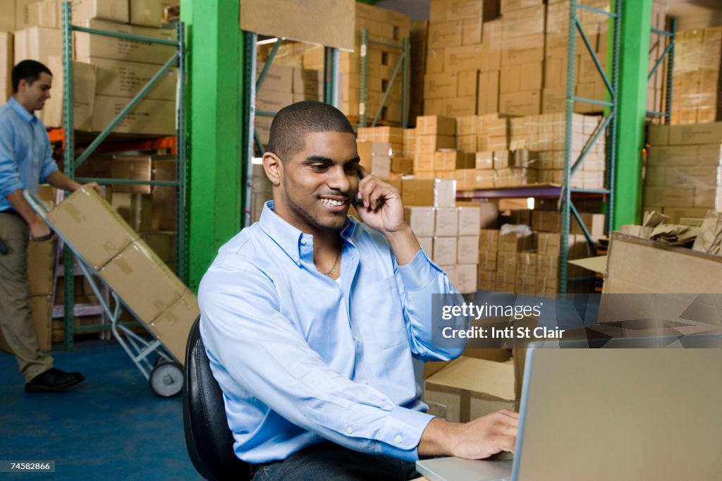 Warehouse worker talking on mobile phone, using laptop, another worker pushing hand truck with boxes in background