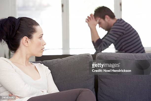 young woman sitting on sofa, looking at distressed man at table - problema di relazione foto e immagini stock