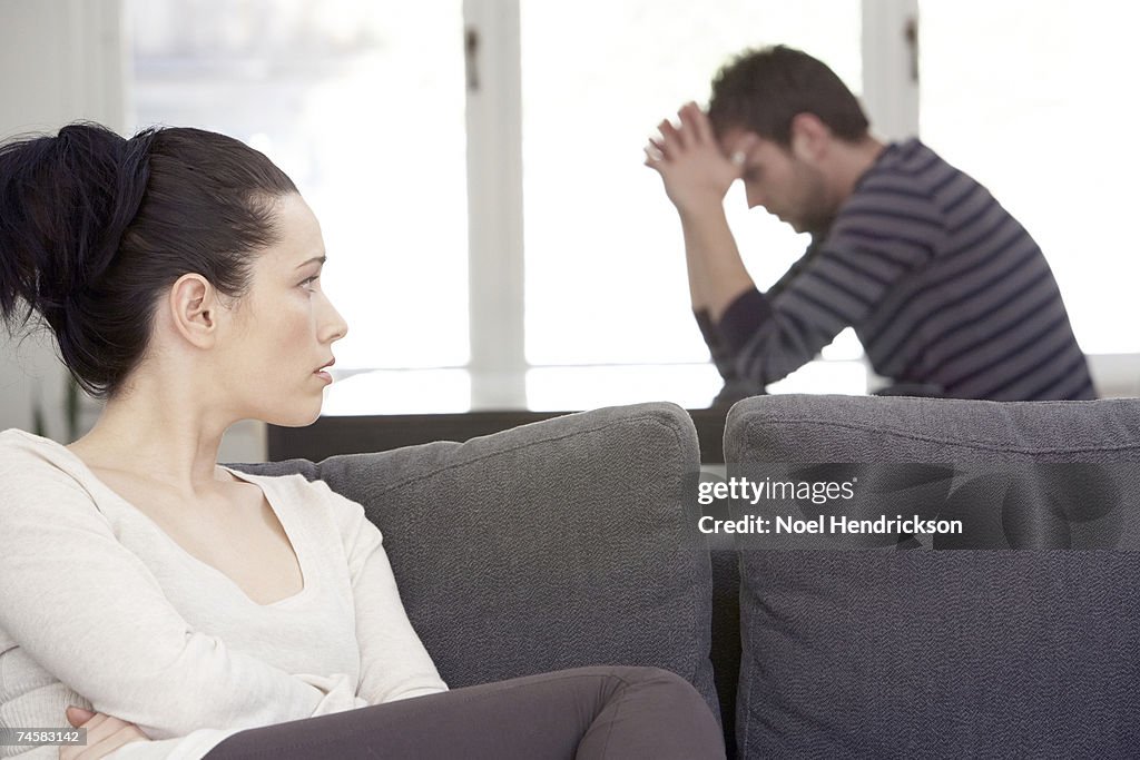 Young woman sitting on sofa, looking at distressed man at table