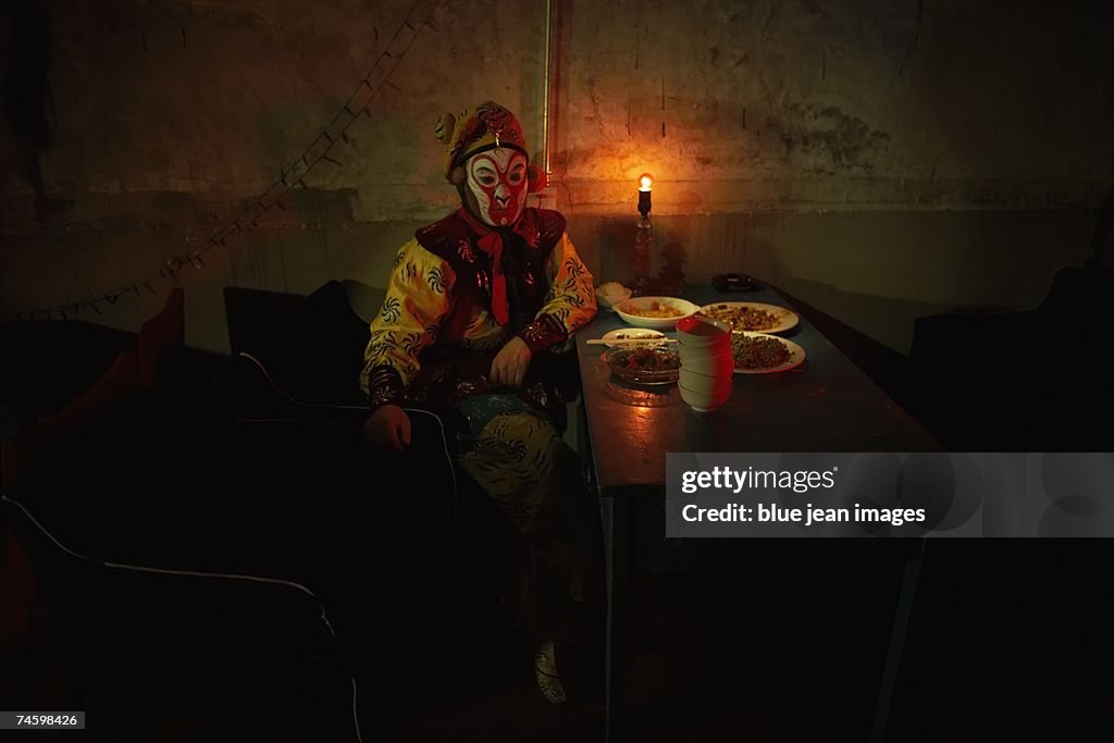 Young actor dressed as a traditional Beijing Opera comedian sits down at a table in front of several dishes of food.