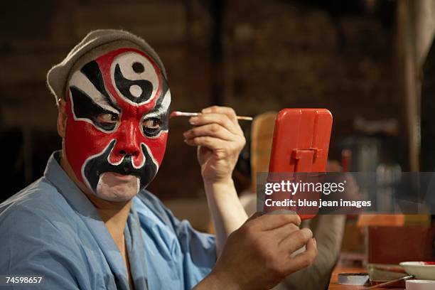 an old actor holds a mirror in front of his face and carefully applies traditional chinese face paint to his eyebrow. - chinese opera stock-fotos und bilder