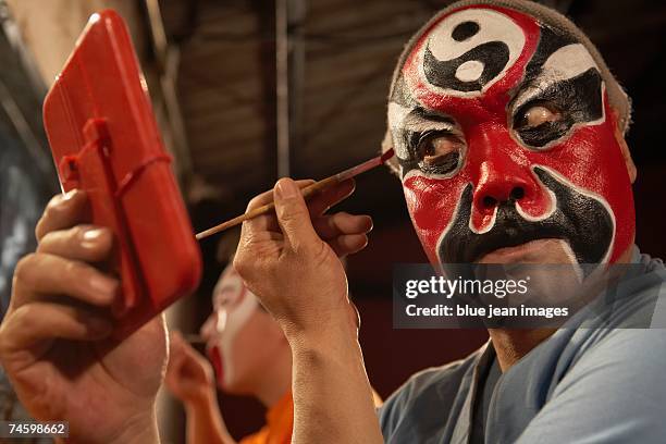 side view of an old actor as he holds a mirror and carefully applies traditional chinese face paint under his eye. - chinese opera stock-fotos und bilder