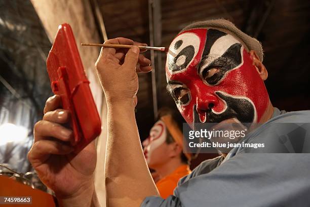 side view of an old actor as he holds a mirror and carefully applies traditional chinese face paint to his forehead. - opera backstage stock pictures, royalty-free photos & images