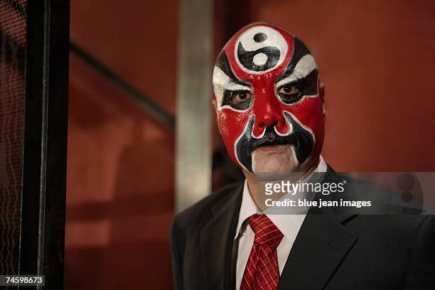 head and shoulders portrait of an old actor posing in front of a stairwell and wearing traditional chinese face paint and a business suit. - chinese mask stockfoto's en -beelden