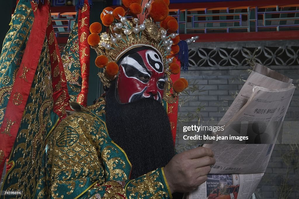 An actor dressed as a traditional Beijing Opera Army General reads the newspaper in an outdoor pavilion.