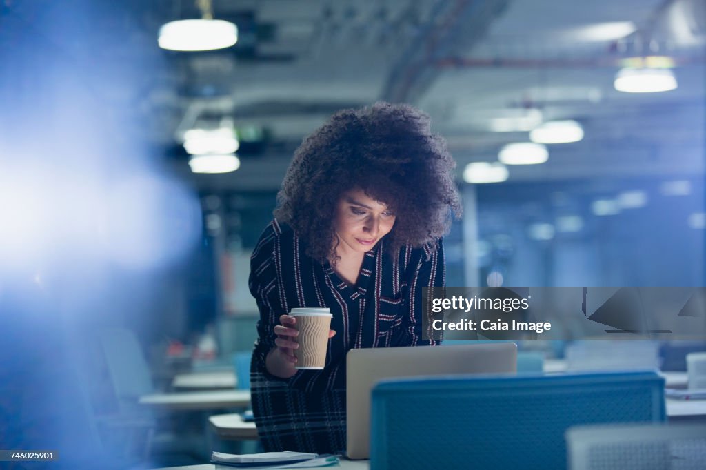 Businesswoman working late at laptop, drinking coffee in dark office