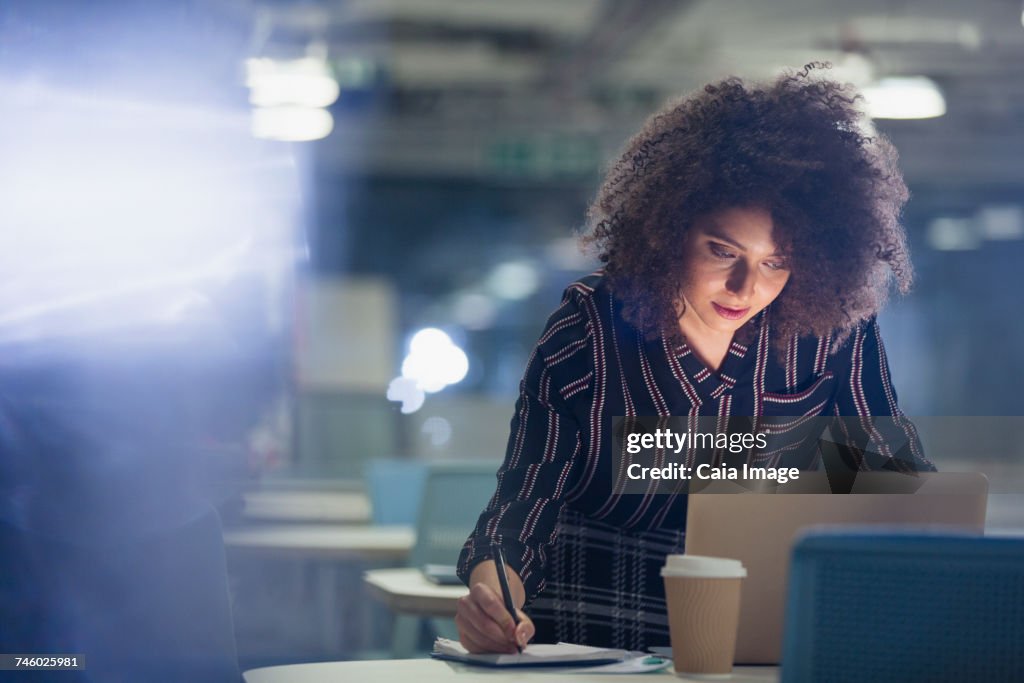 Focused businesswoman working late at laptop, taking notes in dark office
