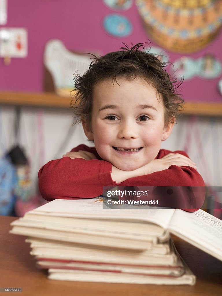 Boy (4-5) sitting at desk and leaning on stack of books, portrait, close-up