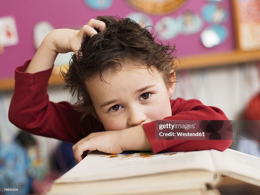 Boy (4-5) sitting at desk with book, hand in hair, portrait, close-up