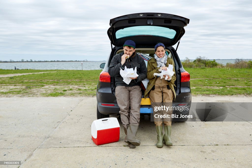 Young couple sitting in open car boot, eating food from paper