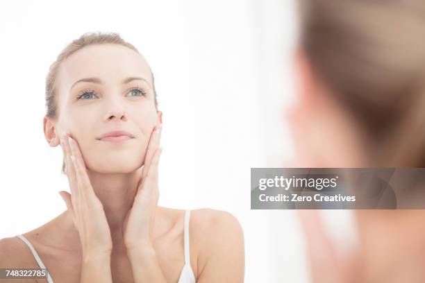 young woman looking in mirror, applying moisturiser to face - gezichtscrème stockfoto's en -beelden