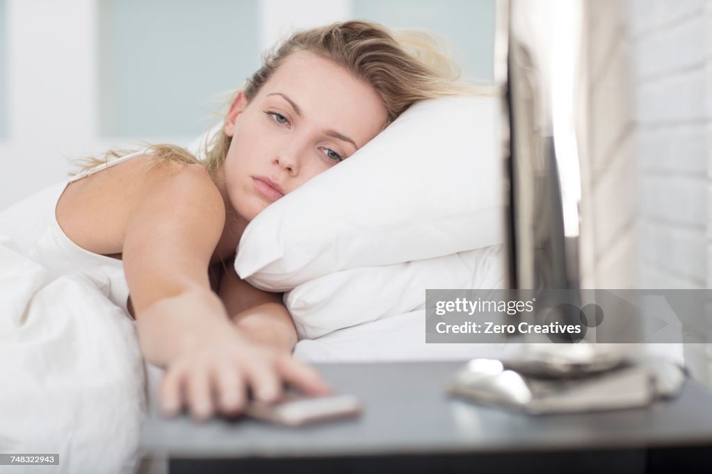 Young woman in bed, reaching for smartphone on bedside table