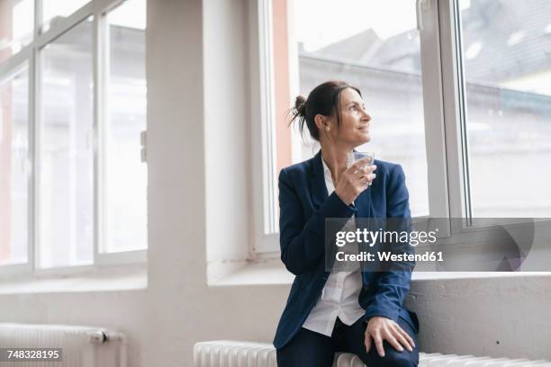 businesswoman with glass of water sitting on radiator in a loft looking through window - drinking water glass woman stock-fotos und bilder