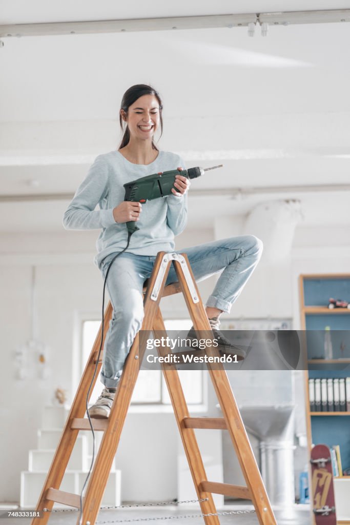 Young woman in her new flat sitting on ladder, holding electric drill